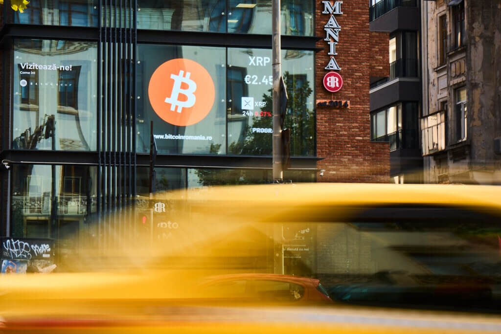 Bitcoin Romania office with large Bitcoin logo on glass facade and blurred yellow taxi passing by