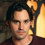 Milo Ventimiglia wearing a maroon cable-knit sweater against a warm, decorative background