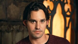 Milo Ventimiglia wearing a maroon cable-knit sweater against a warm, decorative background