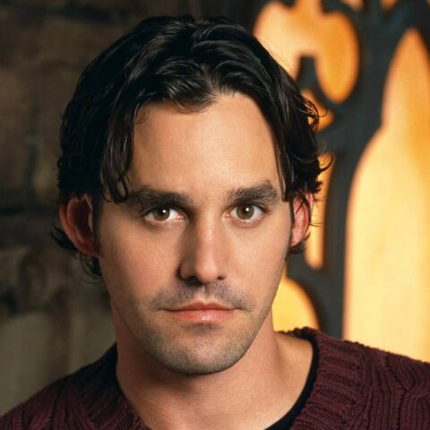 Milo Ventimiglia wearing a maroon cable-knit sweater against a warm, decorative background