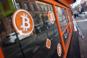 Bitcoin logos on the window of an orange-framed storefront on a city street.