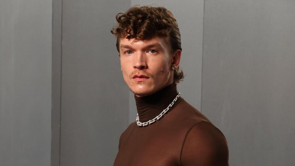 Man wearing a sheer brown turtleneck, black pants, silver chain necklace, and holding a fur coat over his arm