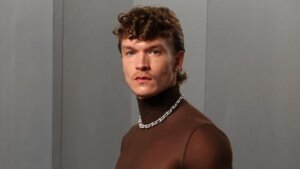 Man wearing a sheer brown turtleneck, black pants, silver chain necklace, and holding a fur coat over his arm