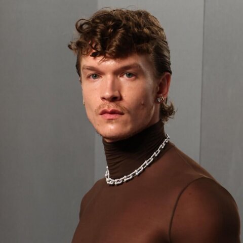 Man wearing a sheer brown turtleneck, black pants, silver chain necklace, and holding a fur coat over his arm