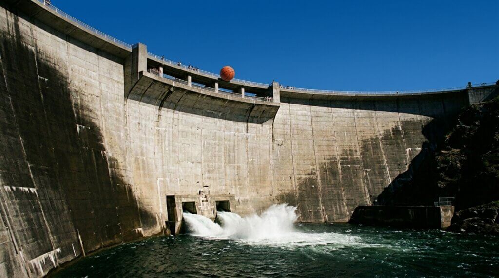 Concrete dam releasing water under a clear blue sky with a basketball digitally added above the dam wall.