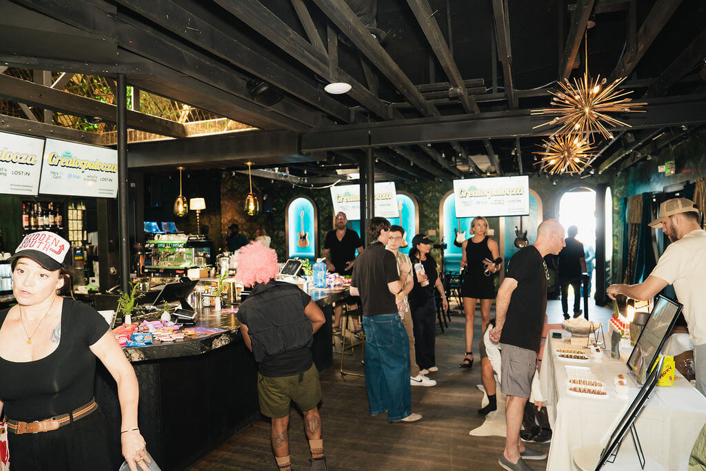 People socializing and browsing tables at an indoor event with "Creatapalooza" signs and guitars on display.