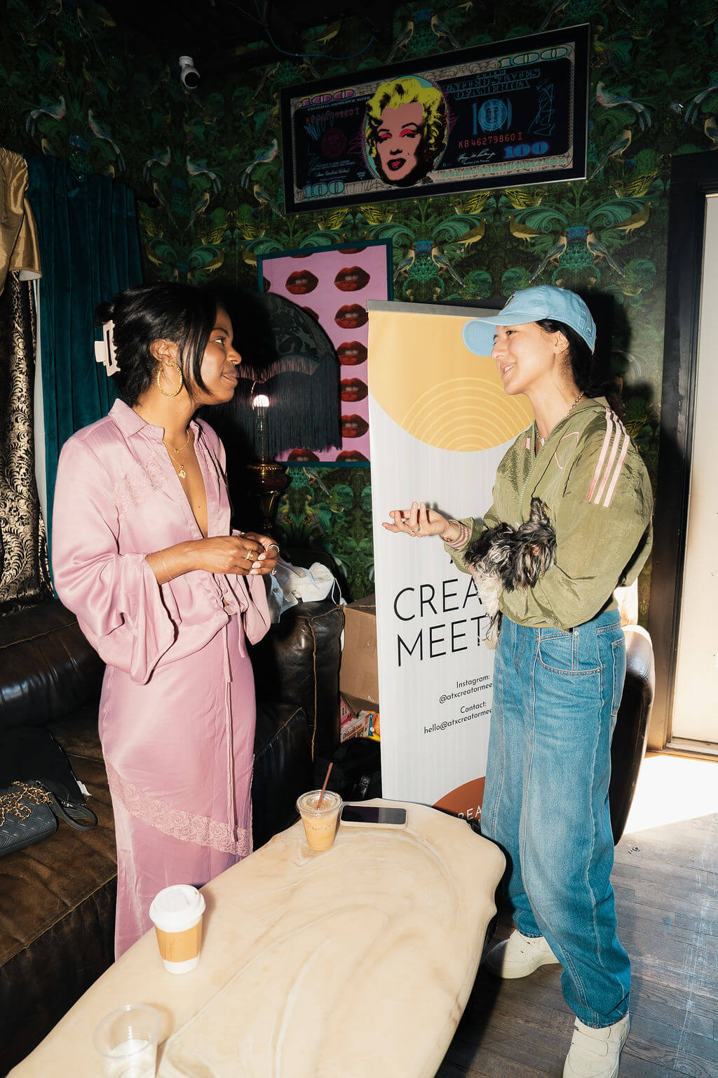 Two women talking indoors near a "CREATOR MEET" banner, one holding a small dog, with Marilyn Monroe art on the wall.