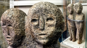 Two ancient stone sculptures of conjoined human figures displayed in a museum case with a chain around them