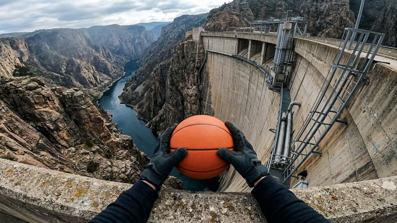 Person wearing black gloves holding a basketball over a large concrete dam with a river and rocky canyon in the background