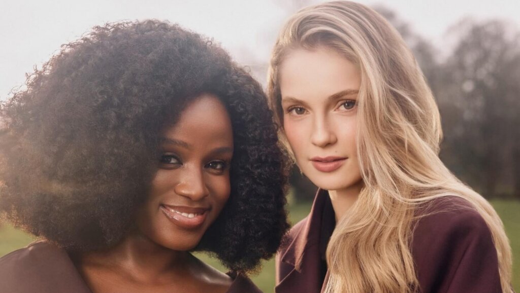Two women wearing dark purple coats posing outdoors with soft natural light and blurred trees in the background