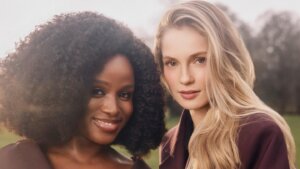 Two women wearing dark purple coats posing outdoors with soft natural light and blurred trees in the background