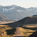 Car driving on a long road through a valley with snow-capped mountains in the background at sunset