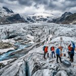 Scientists conducting glacier research with equipment and an orange tent on a snowy glacier under cloudy skies.