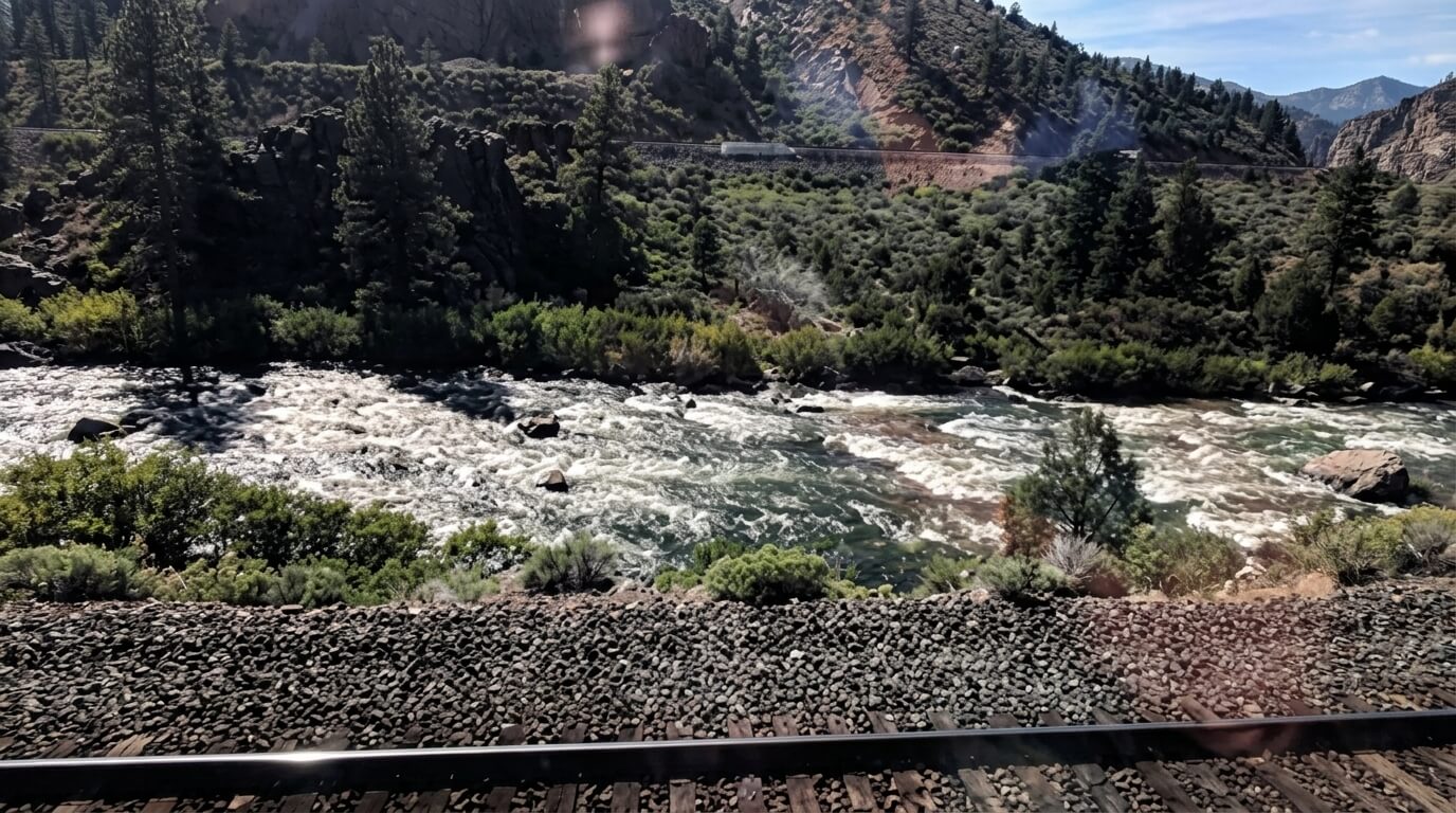 River flowing beside rocky railroad tracks with forested hills and a train in the background