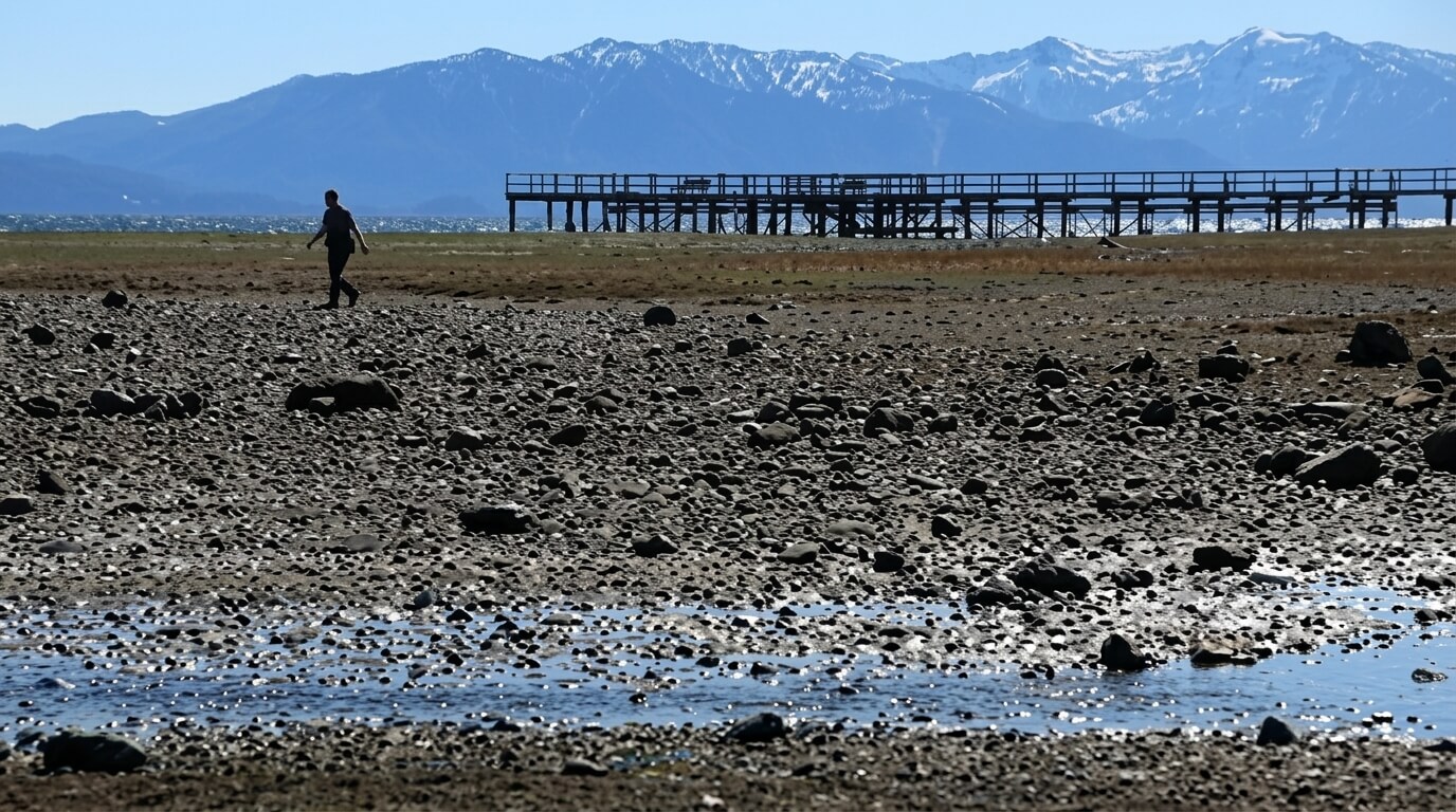 Person walking on rocky shore near wooden pier with snow-capped mountains in the background