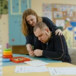 Woman assisting a young man with a developmental disability in a classroom activity with colorful toys and papers.