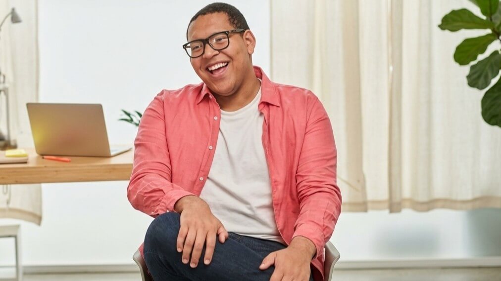 Smiling man wearing glasses and a coral shirt sitting cross-legged in a bright room with a laptop on a desk behind him.