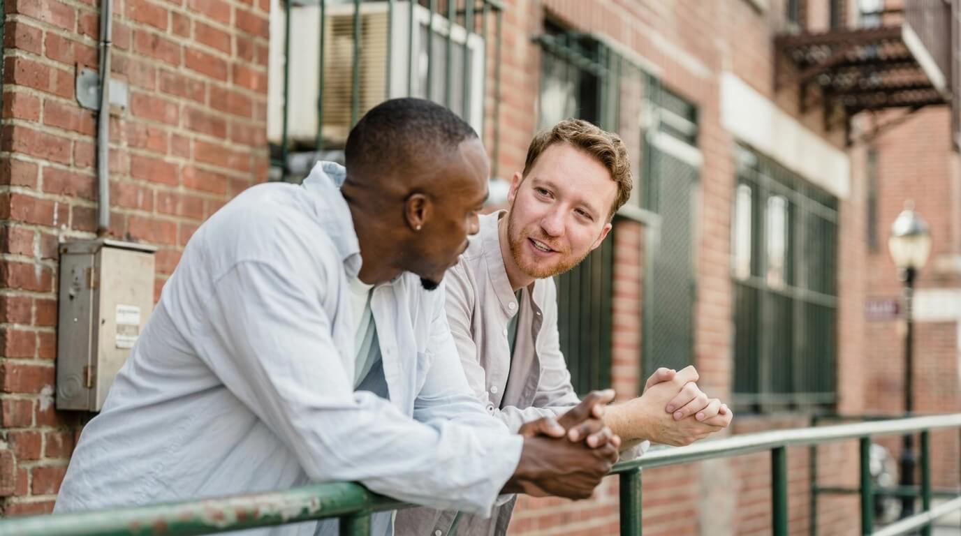 Two men leaning on a green railing having a conversation outside a brick building with barred windows.