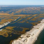 Aerial view of a coastal marshland with waterways, residential houses, and a sandy beach along the ocean.