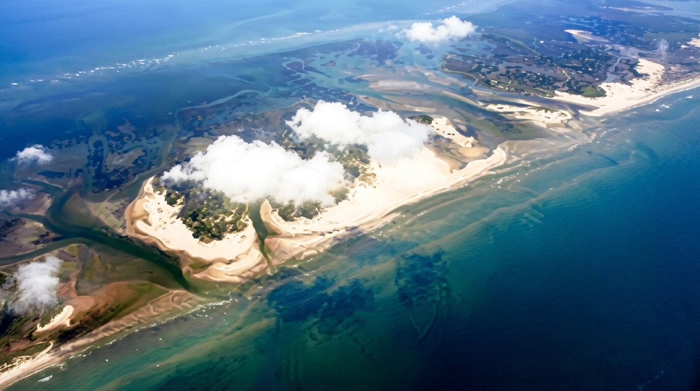 Aerial view of a sandy island with vegetation and clouds surrounded by blue ocean waters.