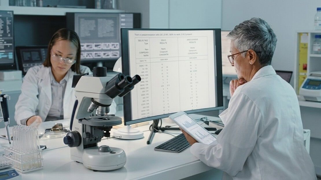 Two scientists in white lab coats analyzing data on a tablet and computer in a laboratory with a microscope.