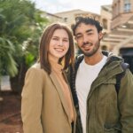 Smiling young couple standing close outdoors near palm trees and urban buildings.