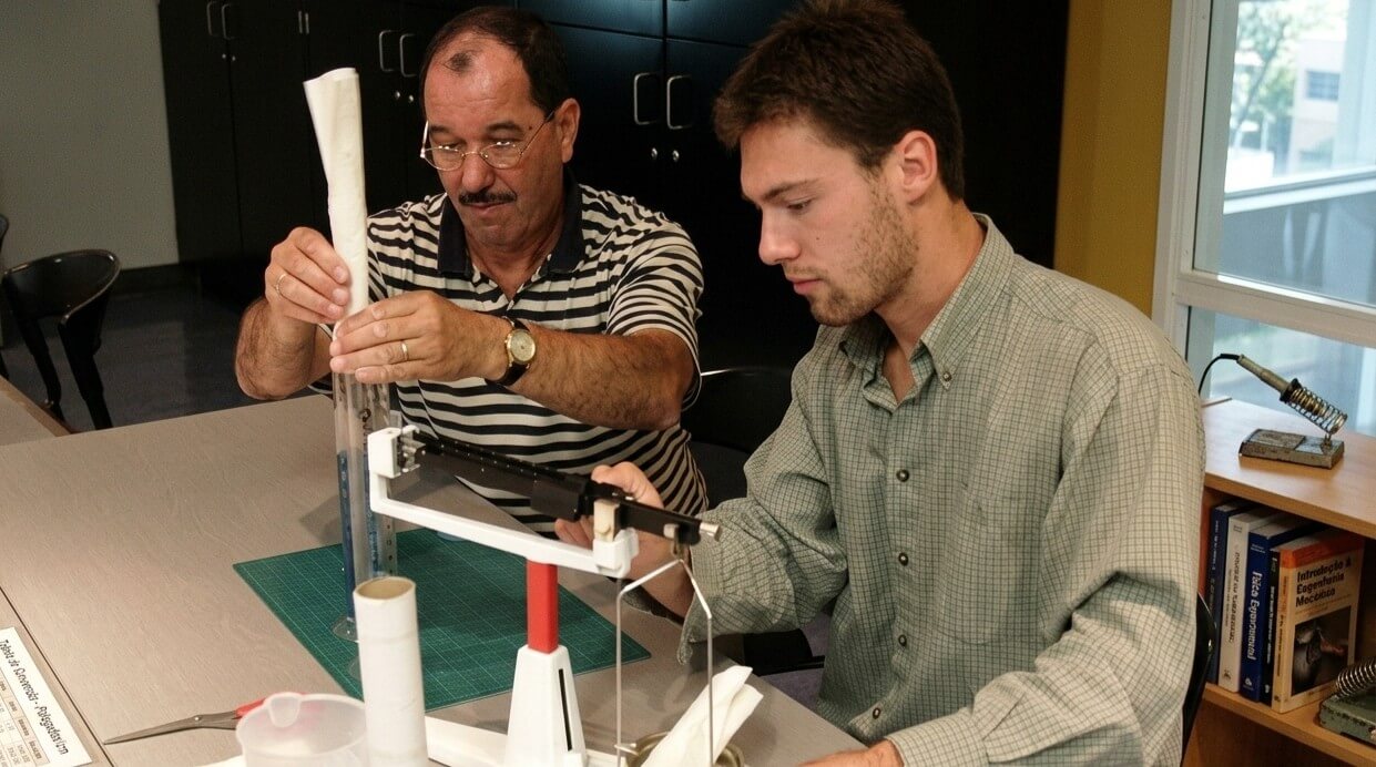 Two men conducting a science experiment with a graduated cylinder and balance scale in a classroom.