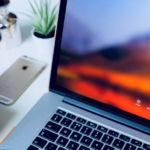 Canon camera, iPhone, and MacBook laptop displaying a login screen on a white desk with a small plant and silver container.