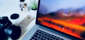Canon camera, iPhone, and MacBook laptop displaying a login screen on a white desk with a small plant and silver container.