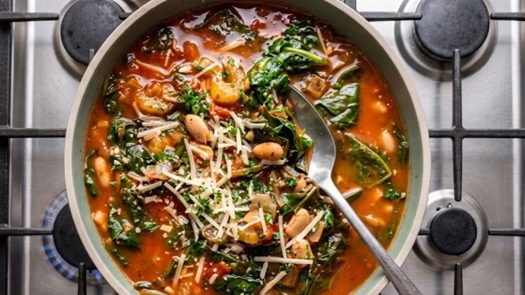 Vegetable and white bean soup with spinach and shredded cheese cooking in a pot on a gas stove.