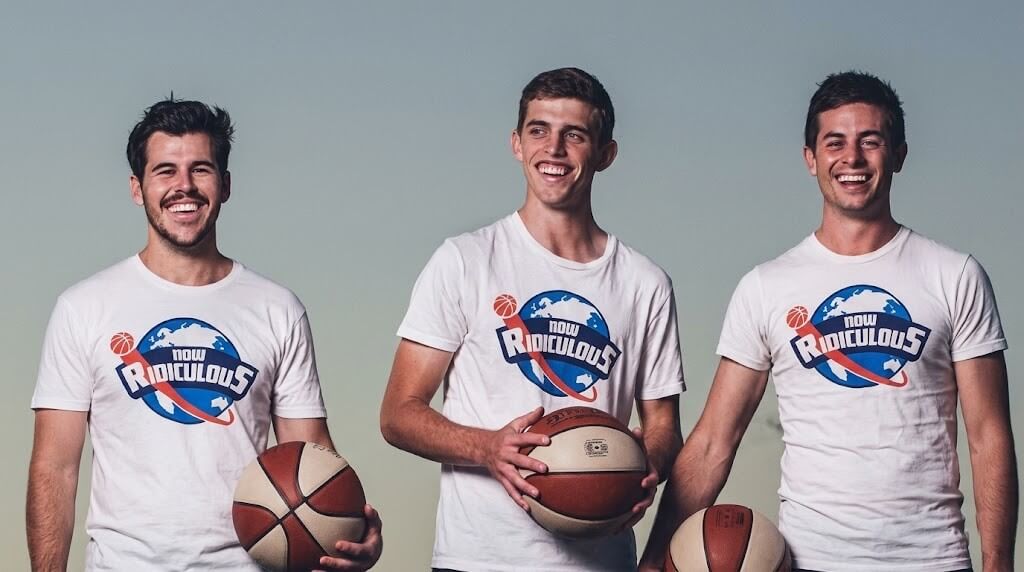 Three smiling men holding basketballs wearing white "Now Ridiculous" t-shirts against a plain background