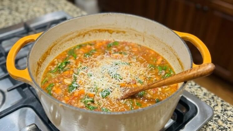 Yellow-handled pot with vegetable soup topped with shredded cheese and spinach on a stovetop