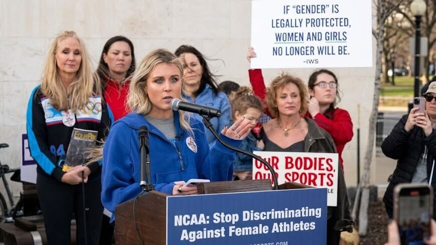 Woman speaking at podium with sign reading "NCAA: Stop Discriminating Against Female Athletes" at protest rally