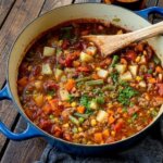 Hearty vegetable and ground meat stew in a blue Dutch oven with a wooden spoon on a wooden table.