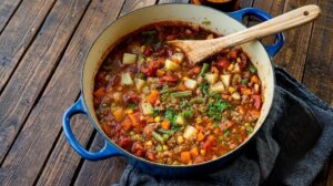 Hearty vegetable and ground meat stew in a blue Dutch oven with a wooden spoon on a wooden table.