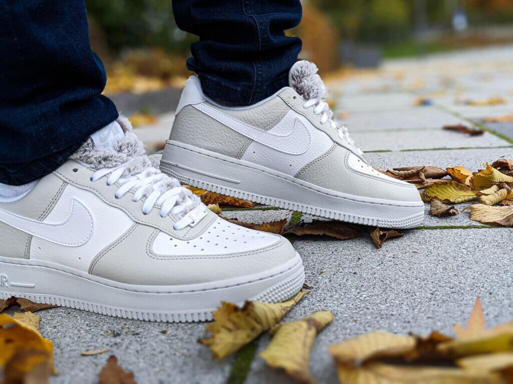 White and gray Nike Air Force 1 sneakers with fuzzy tongue worn with dark jeans on a leaf-covered sidewalk