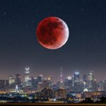Blood moon lunar eclipse over a brightly lit modern city skyline at night with a starry sky.