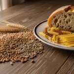 Whole wheat grains with wheat stalks next to a plate holding sliced bread and various uncooked pasta types on a wooden table.