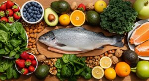 Whole fresh fish on a wooden board surrounded by spinach, strawberries, blueberries, avocados, nuts, citrus fruits, kale, and salmon fillets.