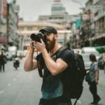 Bearded man wearing a black cap and backpack taking a photo with a Canon camera on a busy city street.