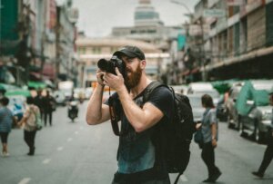 Bearded man wearing a black cap and backpack taking a photo with a Canon camera on a busy city street.