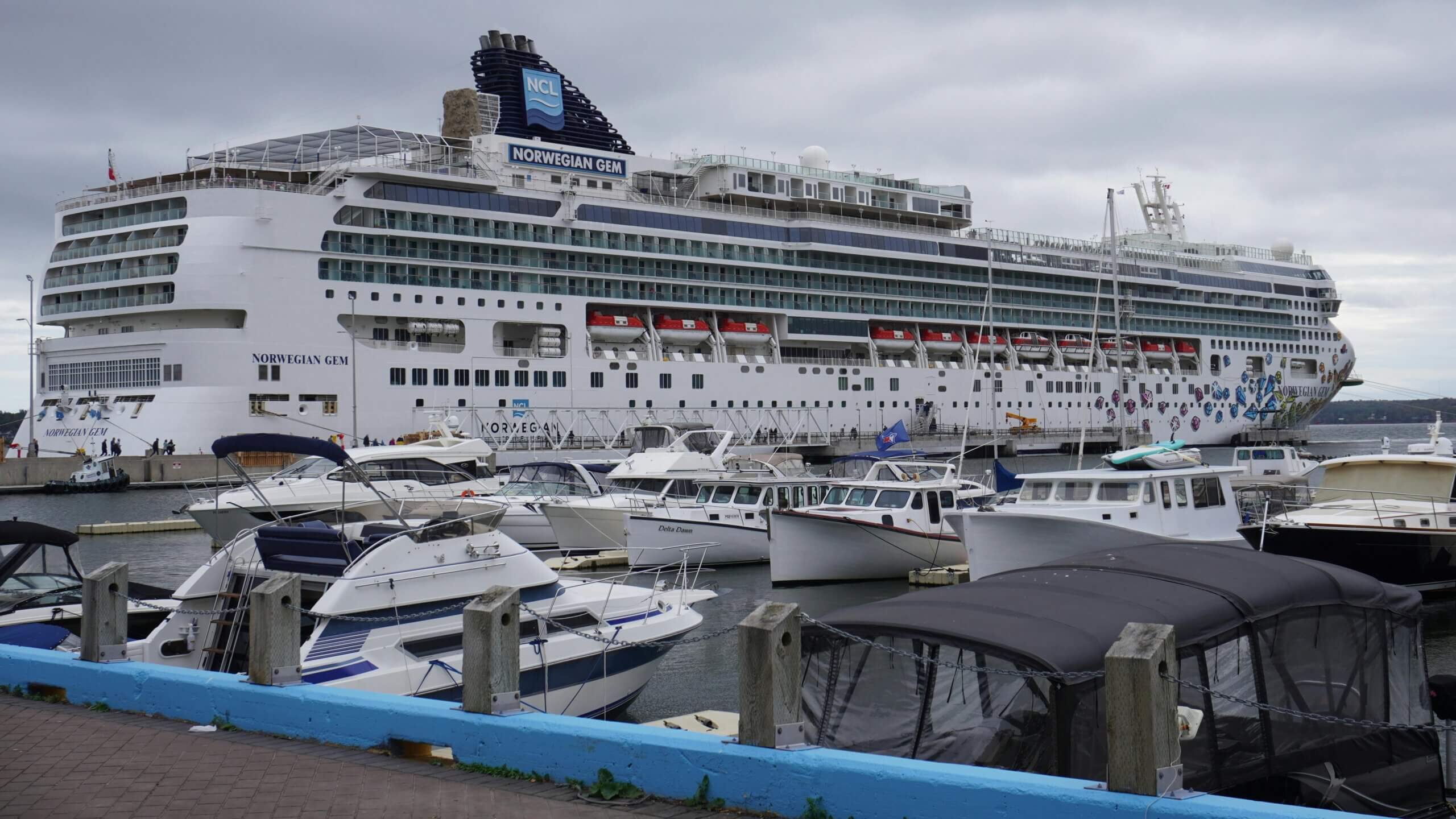 Norwegian Gem cruise ship docked at a marina with smaller boats in the foreground under a cloudy sky.
