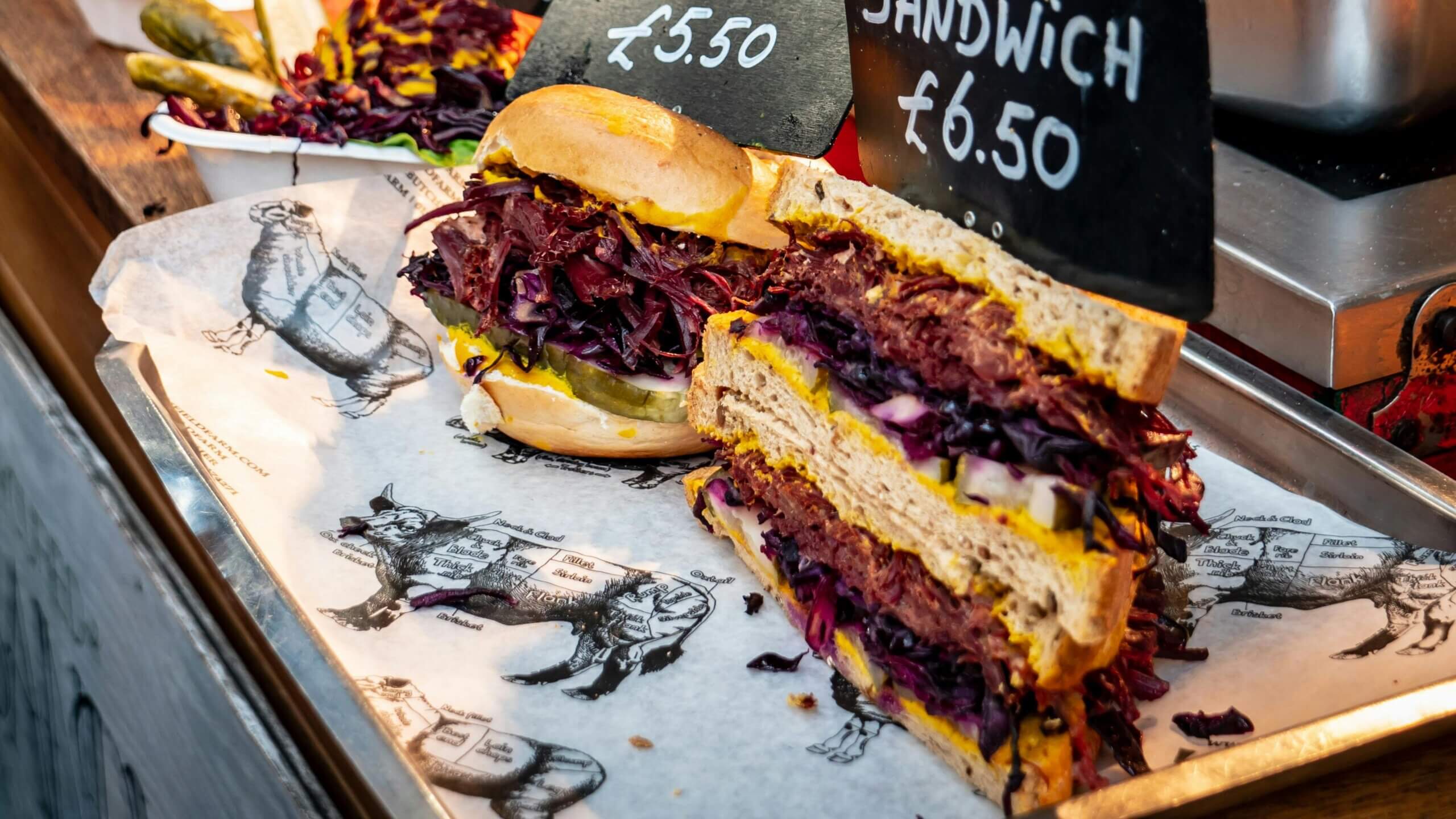 Two sandwiches with pickles, mustard, and red cabbage on a tray with price signs £5.50 and £6.50