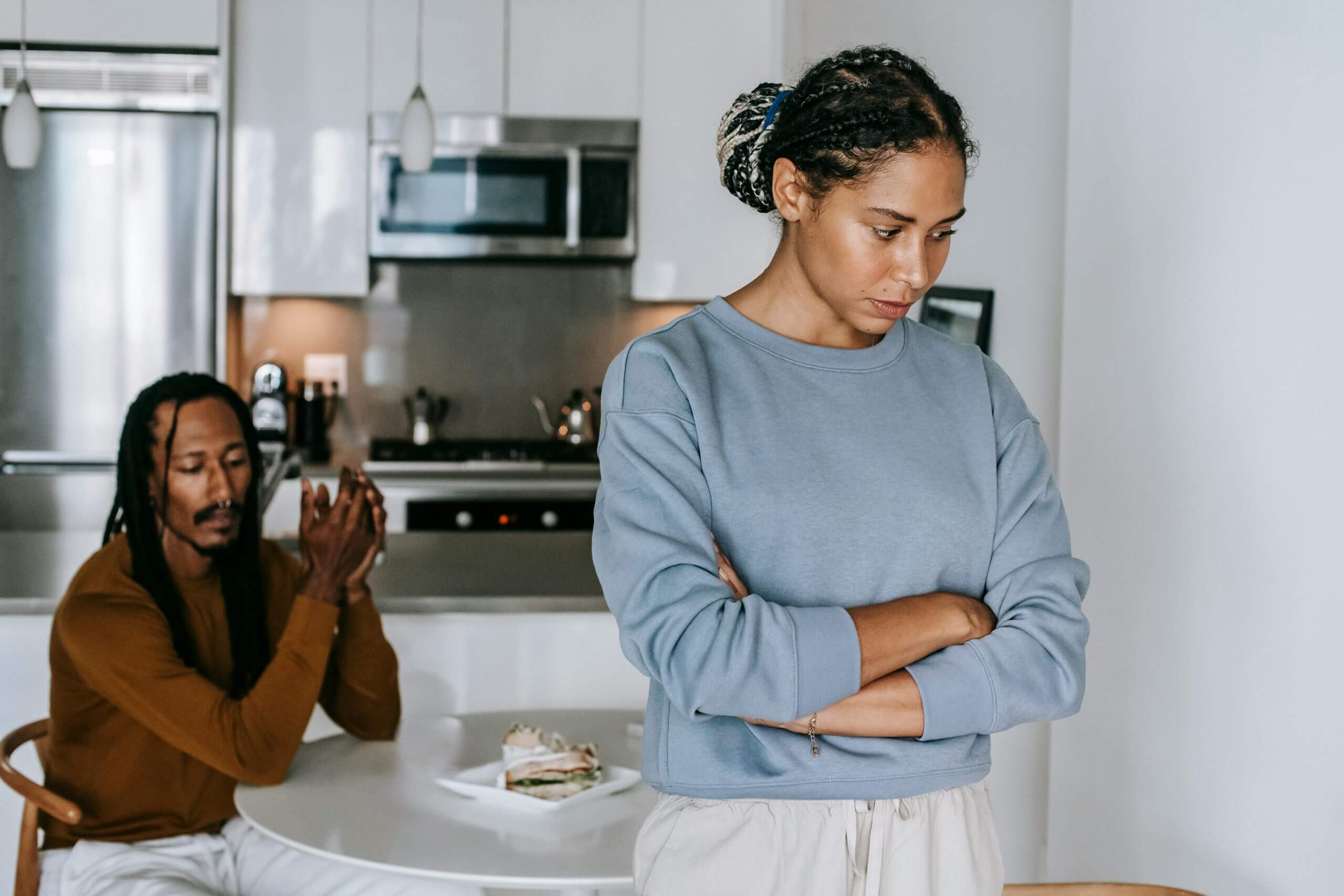 Woman in blue sweatshirt with arms crossed looking down, man in brown shirt sitting at kitchen table behind her with hands clasped