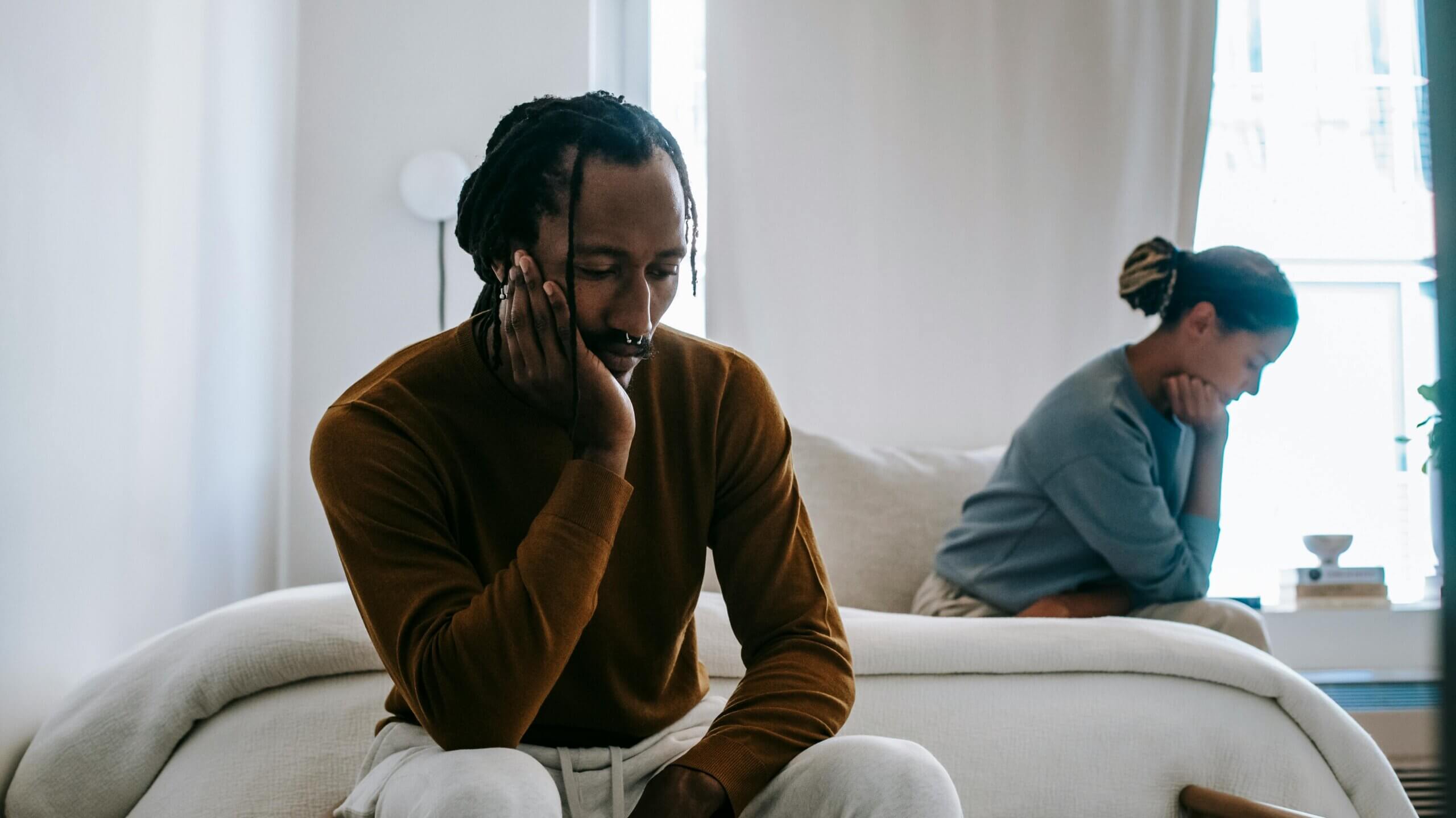 Two people sitting on a bed looking upset and avoiding eye contact in a bright bedroom.