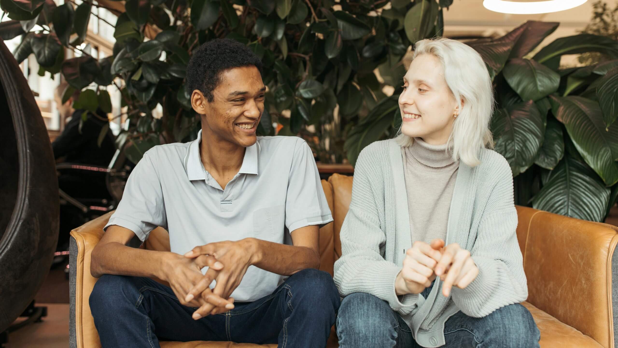 Two young adults smiling and sitting on a brown leather couch with green plants in the background