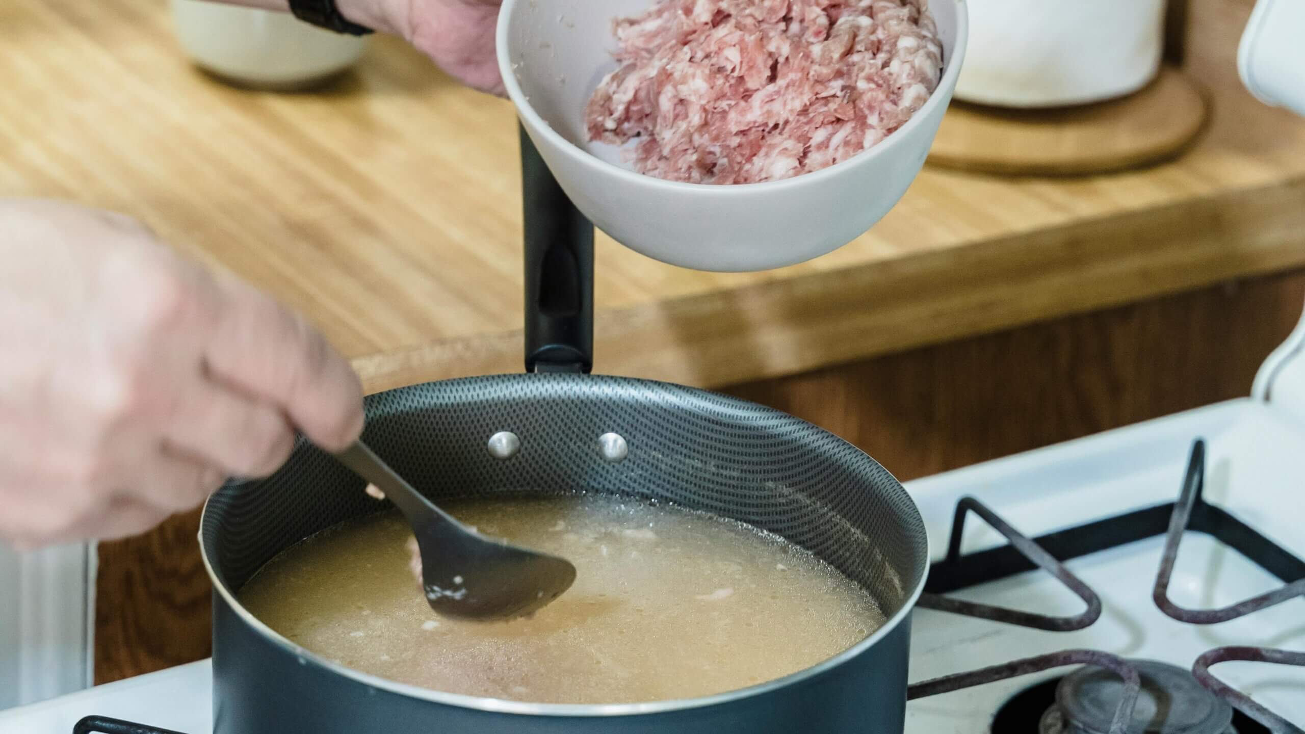 Person adding ground meat from a white bowl into a pot of broth on a gas stove while stirring with a spoon