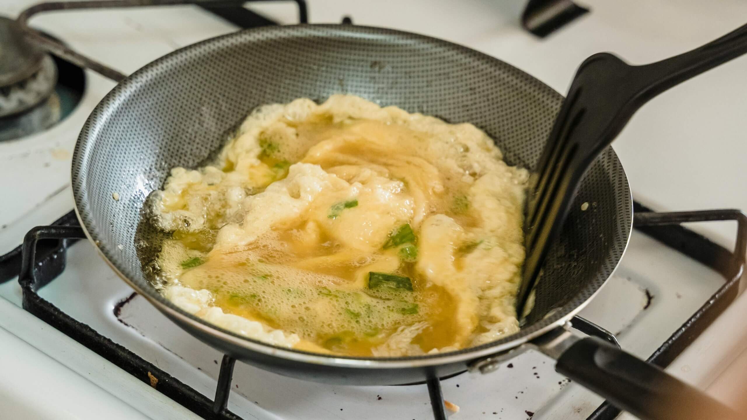 Egg omelette cooking in a non-stick frying pan on a gas stove with a black spatula.