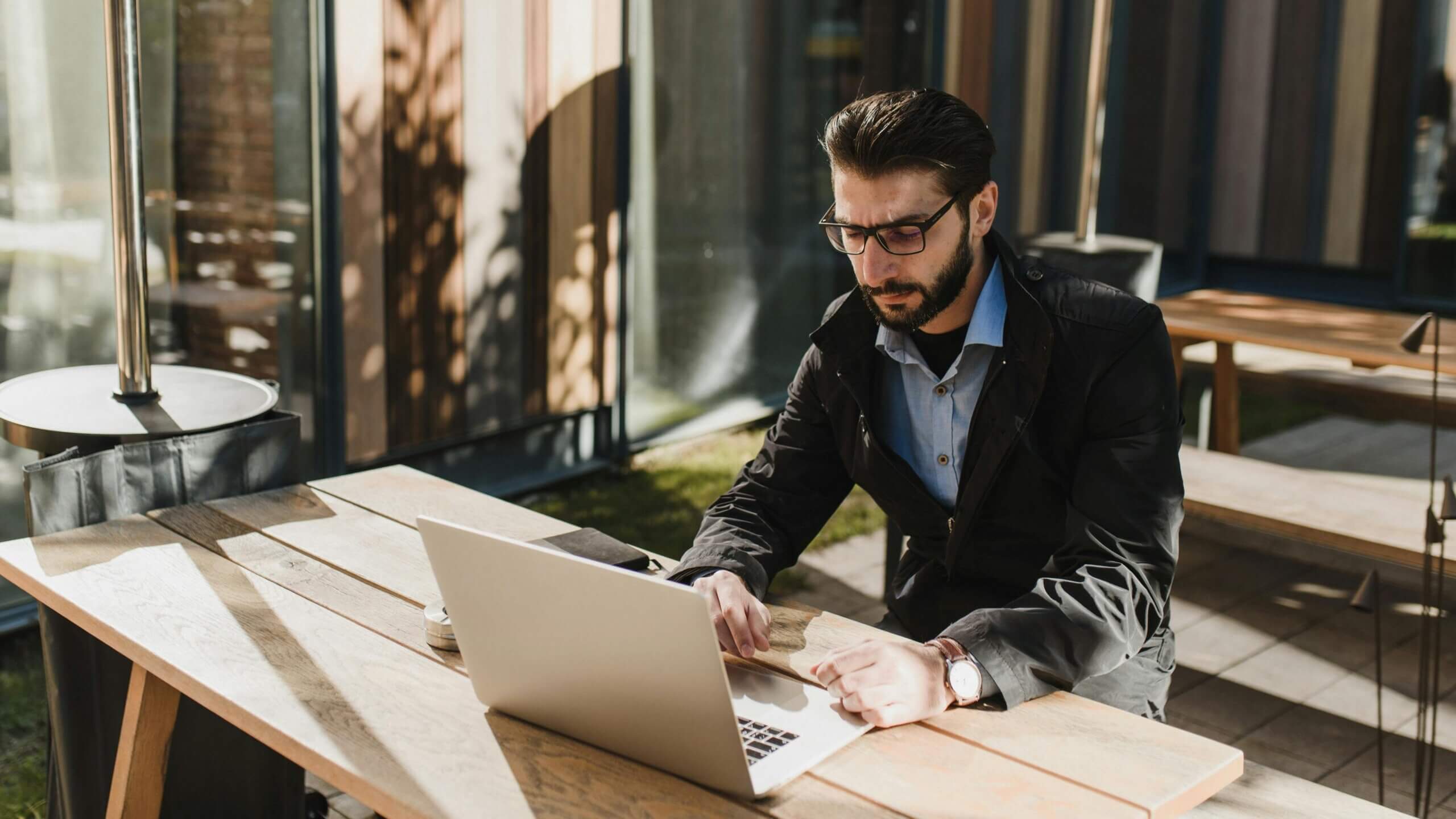 Man with glasses and beard working on a laptop at an outdoor wooden table in daylight