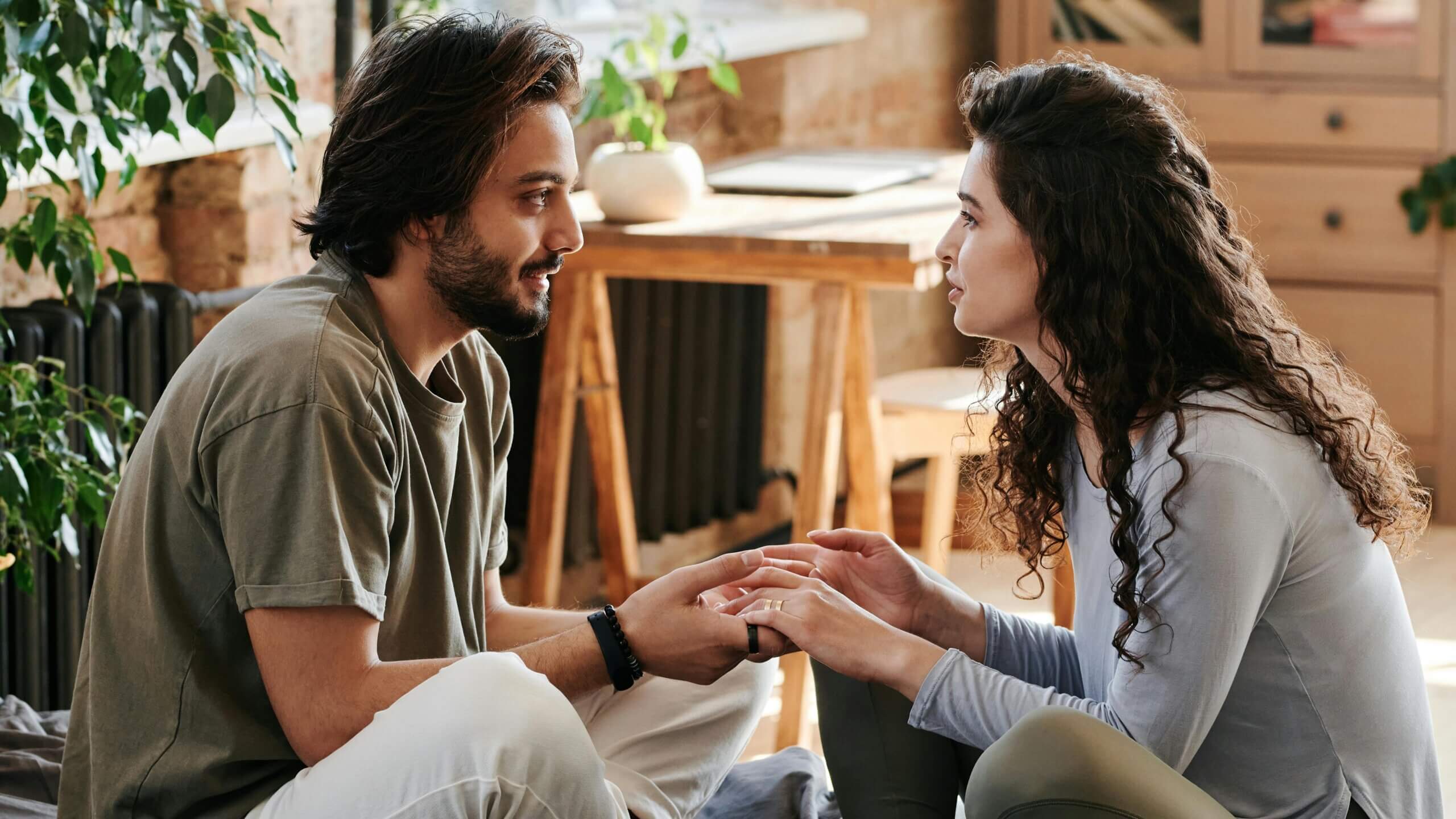 Couple sitting indoors holding hands and looking at each other in a cozy room with plants and wooden furniture.
